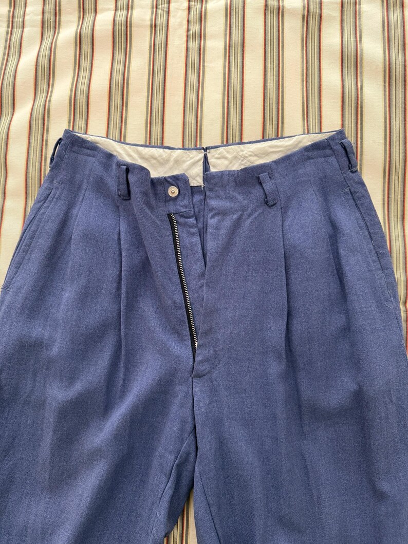 K&ouml;nnte beinhalten: Blaue Hose mit hohem Bund und Rei&szlig;verschluss. Die Hose hat Falten vorne und G&uuml;rtelschlaufen. Der Stoff scheint ein strukturiertes, gewebtes Material zu sein.