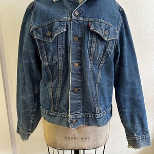 Puede incluir: Una chaqueta de mezclilla azul de lavado medio con cuello clásico, mangas largas y cierre frontal de botones. La prenda incluye dos bolsillos en el pecho con solapas abotonadas, una pequeña etiqueta roja en el bolsillo izquierdo y desgaste visible.