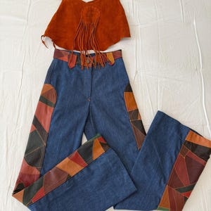 May include: A vintage two-piece outfit featuring a rust-colored suede crop top with fringe and a pair of blue denim bell-bottom pants. The pants have colorful patchwork details on the sides and waistband, creating a retro aesthetic.