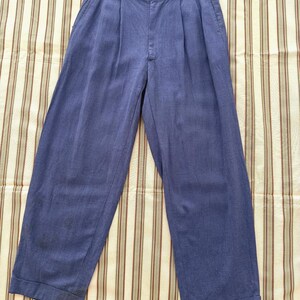 K&ouml;nnte beinhalten: Blaue Hose mit Falten und weitem Bein. Die Hose hat eine hohe Taille und einen Knopf- und Rei&szlig;verschluss. Der Stoff scheint ein leichtes Material zu sein, das f&uuml;r w&auml;rmeres Wetter geeignet ist. Die Hose wird auf einem gestreiften Stoff pr&auml;sentiert.