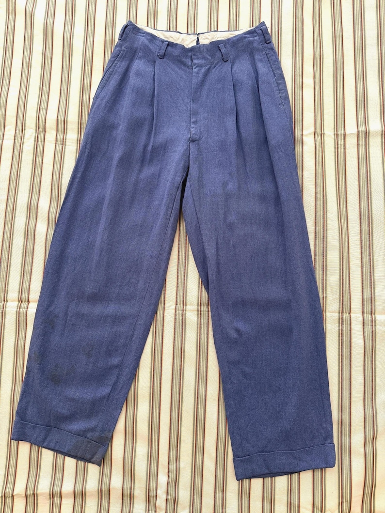 K&ouml;nnte beinhalten: Blaue Hose mit Falten und weitem Bein. Die Hose hat eine hohe Taille und einen Knopf- und Rei&szlig;verschluss. Der Stoff scheint ein leichtes Material zu sein, das f&uuml;r w&auml;rmeres Wetter geeignet ist. Die Hose wird auf einem gestreiften Stoff pr&auml;sentiert.