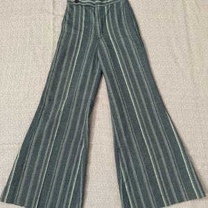 Può includere: Pantaloni a vita alta, a gamba larga, di colore verde acqua scuro con strisce bianche verticali. I pantaloni hanno una chiusura con bottoni e un design svasato, creando un'estetica retrò. Il tessuto sembra essere un materiale tessuto.