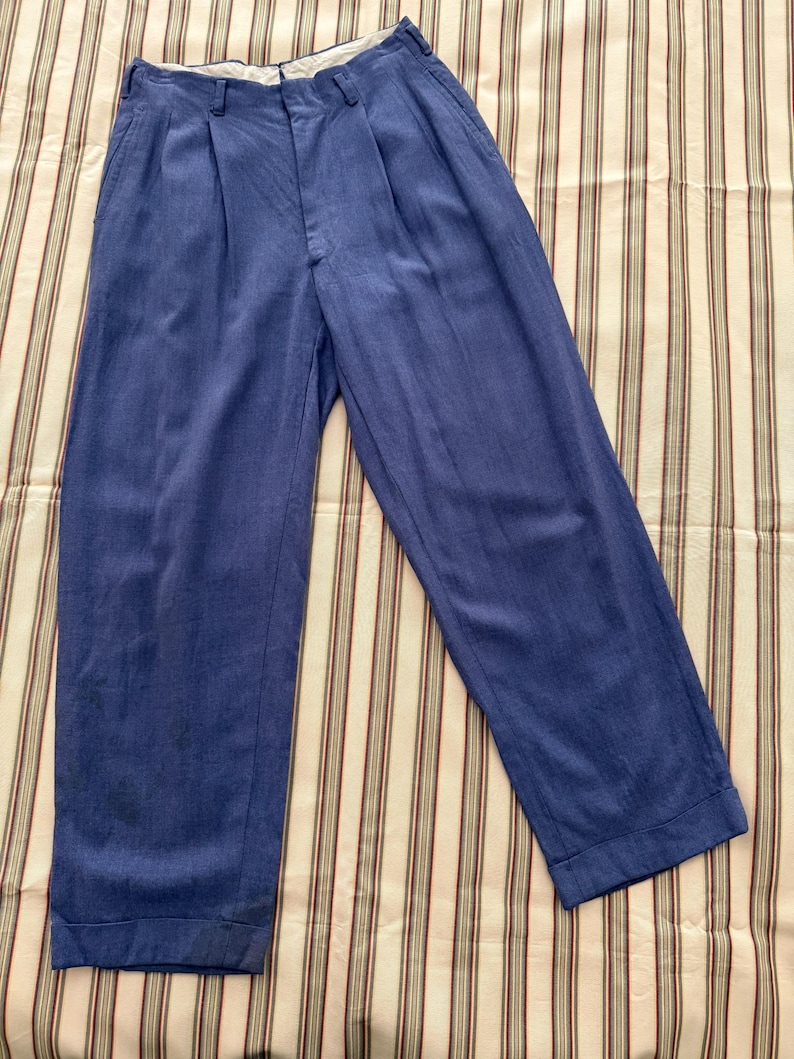 K&ouml;nnte beinhalten: Blaue Hose mit Falten und lockerer Passform. Die Hose ist aus einem weichen Stoff gefertigt und hat ein klassisches Design. Die Hose liegt flach auf einem gestreiften Stoff.