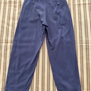 K&ouml;nnte beinhalten: Blaue Hose mit weitem Bein, hohem Bund und zwei Ges&auml;&szlig;taschen mit Kn&ouml;pfen. Die Hose aus weichem Stoff hat einen umgeschlagenen Saum. Pr&auml;sentiert auf einem gestreiften Stoff.