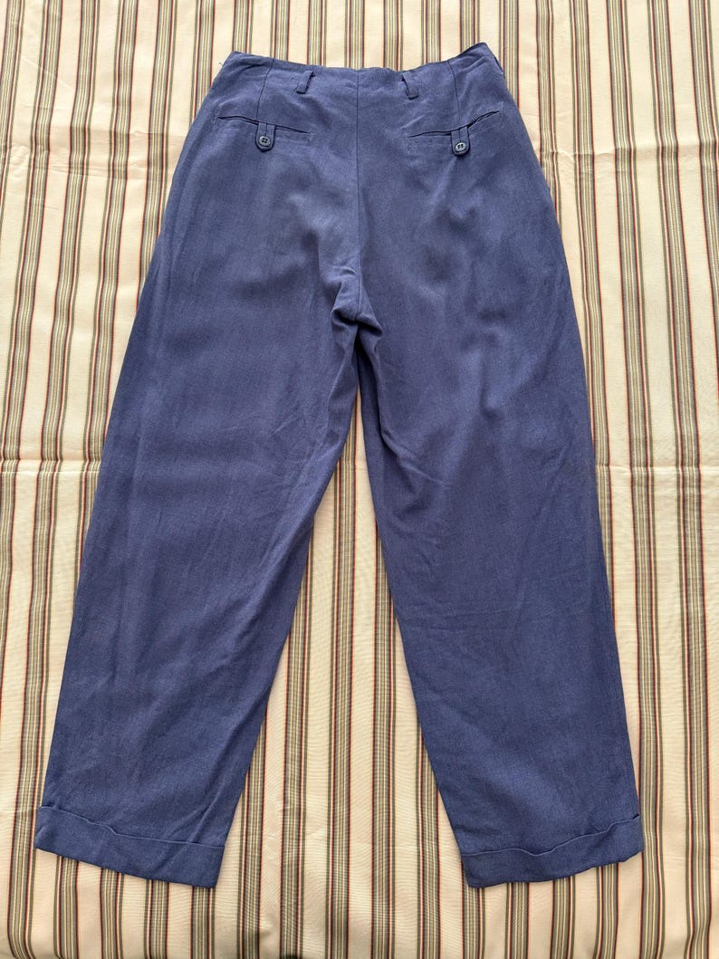 K&ouml;nnte beinhalten: Blaue Hose mit weitem Bein, hohem Bund und zwei Ges&auml;&szlig;taschen mit Kn&ouml;pfen. Die Hose aus weichem Stoff hat einen umgeschlagenen Saum. Pr&auml;sentiert auf einem gestreiften Stoff.