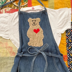 May include: A denim overall dress with a bear applique and a red heart. The dress is paired with a white t-shirt. The bear applique is tan with a red heart. The dress has a halter tie and a waist tie.