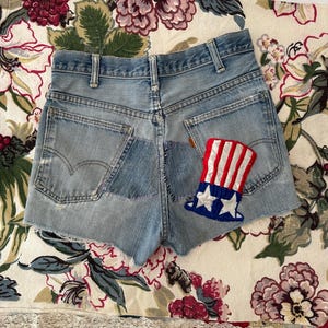 May include: Light blue denim shorts with a distressed look. The shorts feature a patchwork design and a red, white, and blue Uncle Sam hat patch. The shorts have a raw hem and classic five-pocket styling.