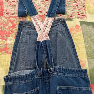 Könnte beinhalten: Blaue Jeans-Latzhose mit weißen Point or Brand Hosenträgern. Die Latzhose hat mehrere Taschen und grüne Nähte. Die Hosenträger kreuzen sich am Rücken und sind mit Metallbeschlägen an der Latzhose befestigt.