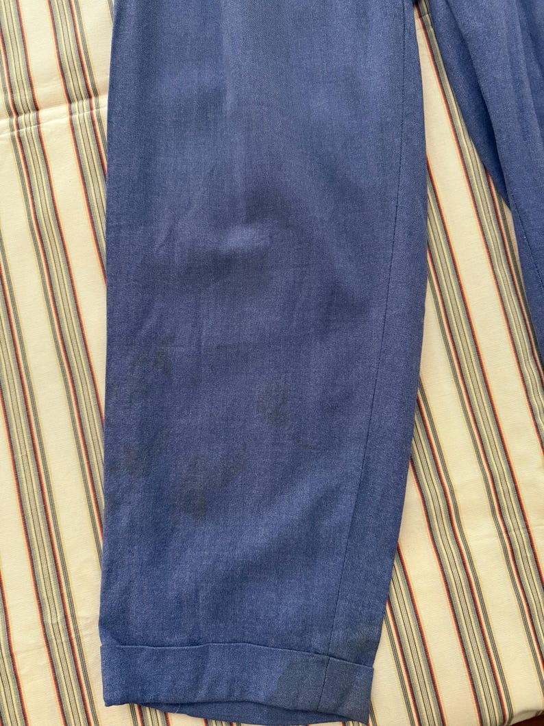 K&ouml;nnte beinhalten: Nahaufnahme einer blauen Hose mit umgeschlagenem Saum. Der Stoff weist einige dunkle Flecken auf und scheint ein mittelschweres Material zu sein. Die Hose liegt auf einem gestreiften Stoff.