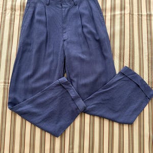K&ouml;nnte beinhalten: Blaue Hose mit Falten vorne und umgeschlagenen S&auml;umen. Die Hose ist aus einem einfarbigen blauen Stoff gefertigt und liegt auf einem gestreiften Hintergrund. Die Hose hat einen Knopf- und Rei&szlig;verschluss.