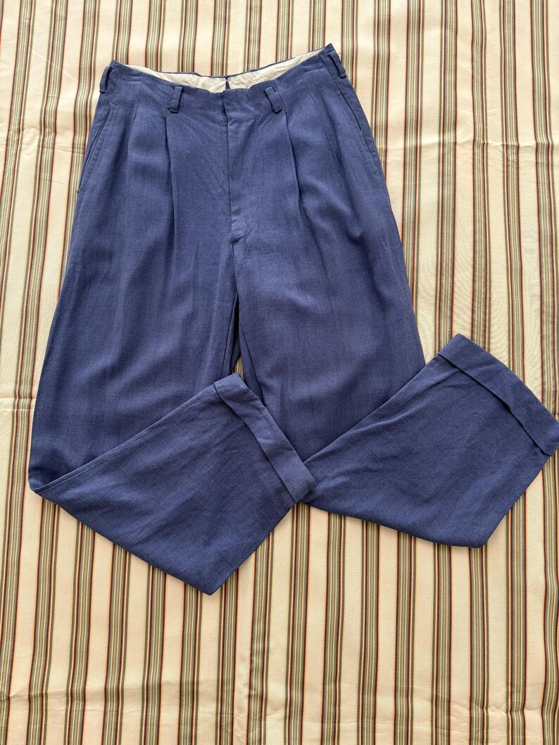K&ouml;nnte beinhalten: Blaue Hose mit Falten vorne und umgeschlagenen S&auml;umen. Die Hose ist aus einem einfarbigen blauen Stoff gefertigt und liegt auf einem gestreiften Hintergrund. Die Hose hat einen Knopf- und Rei&szlig;verschluss.
