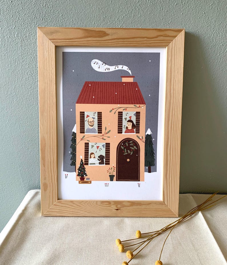 May include: A framed illustration of a house with a red roof and snow on the ground. The house has windows with people looking out, a Christmas tree, and a dog. The illustration is in a light wood frame.