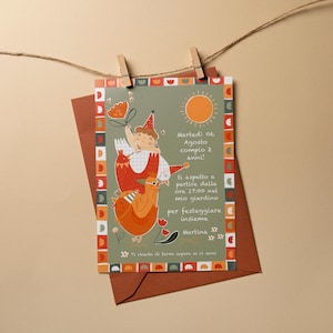 May include: A birthday invitation card with a whimsical illustration of a child riding a bird, with text in Italian. The card is held by a wooden clip on a string, with a brown envelope behind it. The card has a green background and orange accents.