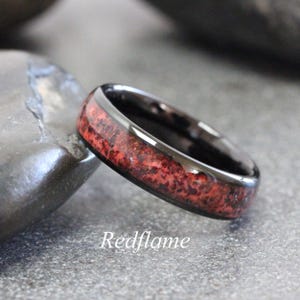 Può includere: Un anello nero con un intarsio rosso strutturato. L'anello è posto su uno sfondo grigio. Il testo "Redflame" è scritto sulla superficie.