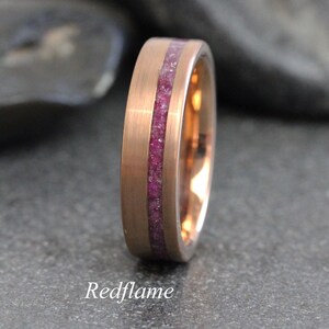 Könnte beinhalten: Roségoldener Ring mit einem zentralen Band aus zerstoßenen, tiefrosa Edelsteinen. Der Ring hat eine gebürstete Metalloberfläche und das Wort "Redflame" ist unten aufgedruckt.