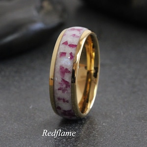 May include: A gold-coloured ring featuring a band of white material inlaid with red fragments. The ring has a polished finish, and the word "Redflame" is printed on the bottom.