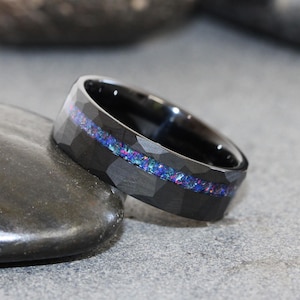 Puede incluir: Un anillo negro con una superficie exterior martillada y facetada y un interior liso y pulido. Una incrustación horizontal central recorre la circunferencia de la banda, presentando un patrón iridiscente de ópalo triturado con motas azules, moradas y rosas.