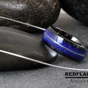 May include: A black ceramic ring with a blue lapis lazuli inlay. The ring is set on a gray stone surface. REDFLAME Jewelry is printed on the surface.
