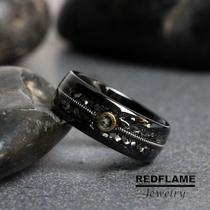 Op de afbeelding: Een zwarte ring met een uniek ontwerp. De ring heeft een gitaarsnaar en een goudkleurig accent. De band is zwart met een textuurinleg. De ring wordt gepresenteerd op een grijs oppervlak.