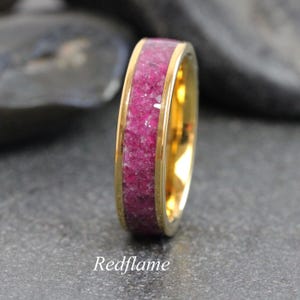 May include: A gold-coloured ring featuring a band of crushed, vibrant pink gemstones. The ring has a polished finish and the text "Redflame" is visible at the bottom of the image. The ring is set against a dark background.