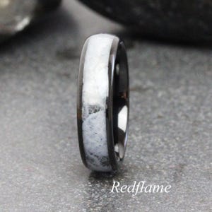 May include: A black ring with a white and gray stone inlay. The ring has a polished finish and a rounded profile. The text "Redflame" is at the bottom of the image.
