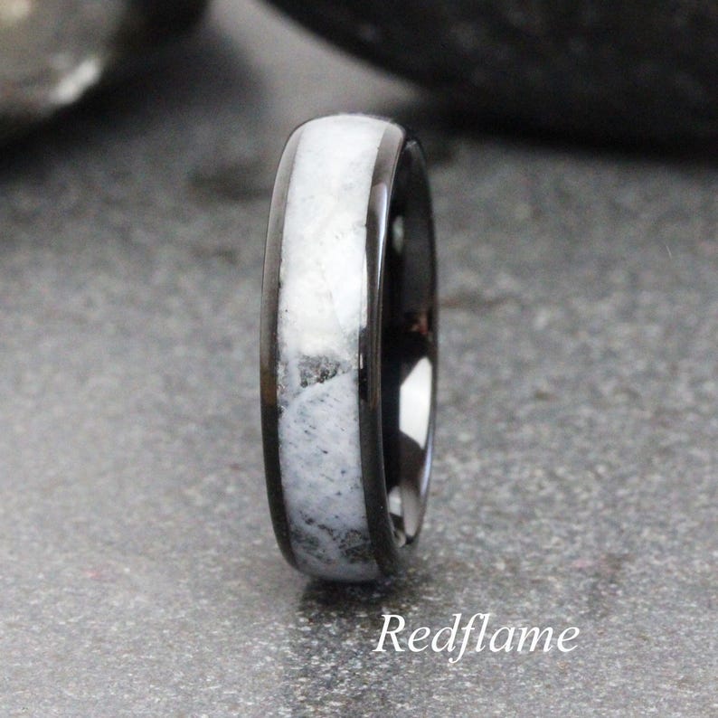 May include: A black ring with a white and gray stone inlay. The ring has a polished finish and a rounded profile. The text "Redflame" is at the bottom of the image.
