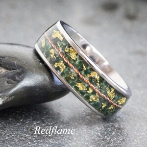 Puede incluir: Un anillo plateado con una incrustación de piedra verde, detalles de pan de oro y un alambre de cobre en el centro. El anillo está sobre un fondo gris. La palabra "Redflame" está en la parte inferior.