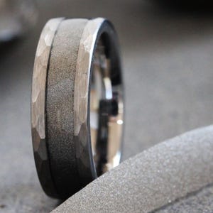 Puede incluir: Un anillo metálico con una banda central ancha de color gris oscuro con acabado arenado texturizado, flanqueado por dos bordes plateados pulidos que muestran un patrón martelado y facetado a lo largo de la circunferencia exterior.