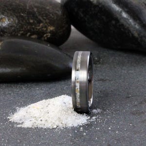 May include: A silver wedding band with a thin white inlay. The band is sitting on a pile of white sand.