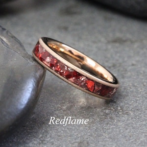 Puede incluir: Un anillo de oro rosa con una banda con incrustaciones de piedras preciosas rojas vibrantes. El diseño del anillo presenta una engaste al ras de las piedras, creando una superficie continua y texturizada. La palabra "Redflame" está escrita en cursiva.