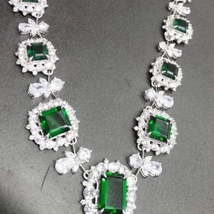 May include: A silver necklace with emerald green gemstones set in square frames. The necklace is adorned with smaller clear gemstones.