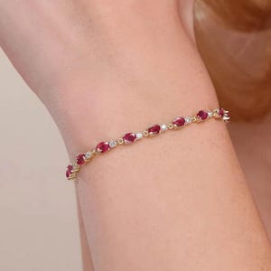 May include: A gold bracelet featuring oval-cut red gemstones and small, round white gemstones. The bracelet is a delicate piece of jewelry, with the gemstones set closely together. The bracelet is worn on a person's wrist.