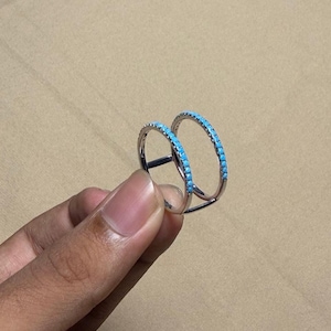 May include: A silver double-band ring with a row of small, bright turquoise stones set along the top of each band. The ring is held up against a neutral background.