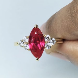 Puede incluir: Un anillo de oro con una gran piedra preciosa roja en forma de marquesa rodeada de pequeñas piedras preciosas blancas.