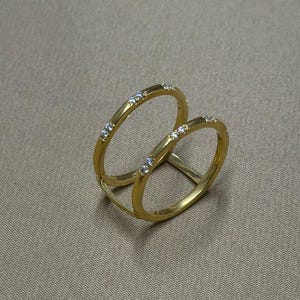 May include: A gold ring featuring two linked hoops, each embellished with tiny, clear gemstones. The ring's design is showcased against a neutral backdrop. A piece of jewellery.