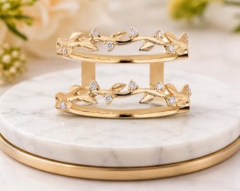 May include: A gold double-band ring featuring two parallel vine-inspired branches. Each branch is adorned with small, polished gold leaves and several round, clear, sparkling stones set at intervals along the delicate, winding metal stems.