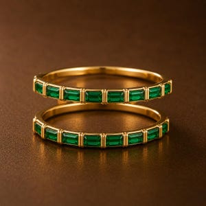 May include: Two gold rings featuring a continuous row of emerald green baguette-cut stones set in a channel setting. The rings have a polished gold band and a uniform design with rectangular green stones encircling the entire circumference.