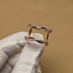 Könnte beinhalten: Roségoldener Ring mit Doppelband-Design. Jedes Band weist ein verdrehtes, seilartiges Muster auf, das mit kleinen, funkelnden, klaren Steinen besetzt ist. Der Ring wird von einer behandschuhten Hand vor neutralem Hintergrund gehalten.