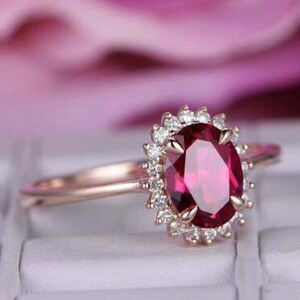 Könnte beinhalten: Ein Verlobungsring aus Roségold mit einem großen ovalen Rubin, der von einem Heiligenschein aus kleineren Diamanten umgeben ist.