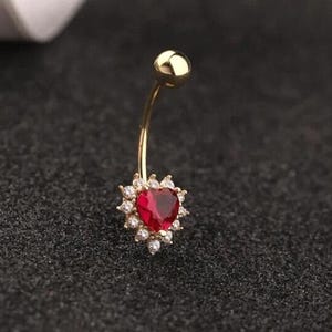 Könnte beinhalten: Goldener Bauchnabelring mit einem roten herzförmigen Stein, der von klaren Strasssteinen umgeben ist.