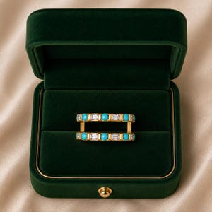 Puede incluir: Dos anillos de oro con piedras turquesas y de talla baguette se exhiben en un joyero de terciopelo verde. Los anillos están paralelos, mostrando las vibrantes piedras azules y transparentes.