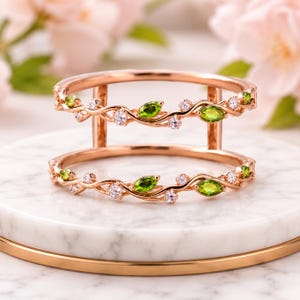 May include: A double-band rose gold ring featuring a vine-like design. The bands are adorned with marquise-cut green peridot gemstones and small, round, clear white stones set along the twisted metal branches that connect the two rings.