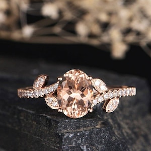 Puede incluir: Anillo de oro rosa con una piedra preciosa ovalada de color melocotón en el centro. La banda está adornada con pequeñas piedras blancas brillantes y detalles en forma de hojas, creando un diseño elegante e inspirado en la naturaleza. El anillo se encuentra sobre un fondo oscuro.