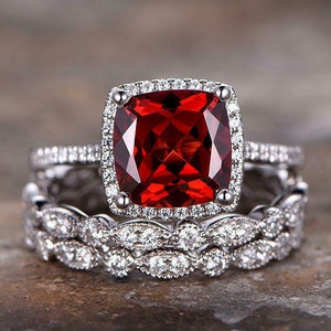 Pode incluir: Um conjunto de anéis de duas peças. O anel principal apresenta uma grande pedra preciosa vermelha escura de corte quadrado, rodeada por um halo de pedras claras menores. A banda também é adornada com pedras menores. Os anéis estão sobre uma superfície neutra.