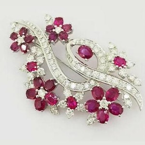 Puede incluir: Broche floral con base de metal plateado. El broche presenta piedras preciosas de color rubí dispuestas en forma de flores, intercaladas con piedras preciosas claras y brillantes. El diseño es elegante y detallado, con una forma fluida.