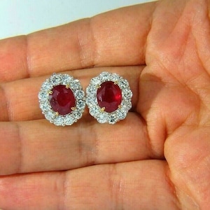 Pode incluir: Um par de brincos ovais de rubi e diamantes. Os rubis são de cor vermelha intensa e são cercados por um halo de diamantes menores.