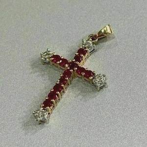 Puede incluir: Un colgante de cruz dorada con piedras rojas en cada brazo. La cruz tiene piedras blancas cuadradas en los extremos. El colgante tiene un pequeño lazo en la parte superior para una cadena.