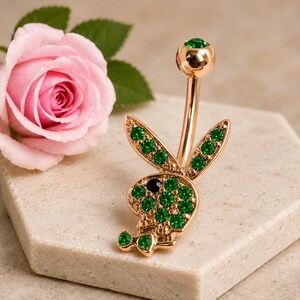 Pode incluir: Um piercing de umbigo em tom de ouro rosa com um pingente em forma de silhueta de coelho incrustado com cristais verdes vibrantes e um olho de cristal preto. A barra é rematada por uma esfera a condizer em tom de ouro rosa com cristal verde.
