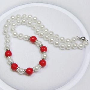 Puede incluir: Un collar de perlas blancas con un brazalete de cuentas rojas y blancas. El collar está hecho de perlas falsas y el brazalete tiene cuentas rojas con diamantes de imitación transparentes.