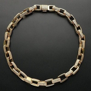 May include: A gold-colored chain necklace with rectangular links, each encrusted with small, sparkling stones. The necklace is laid out on a dark surface, showcasing its design and the way it would lay around the neck.